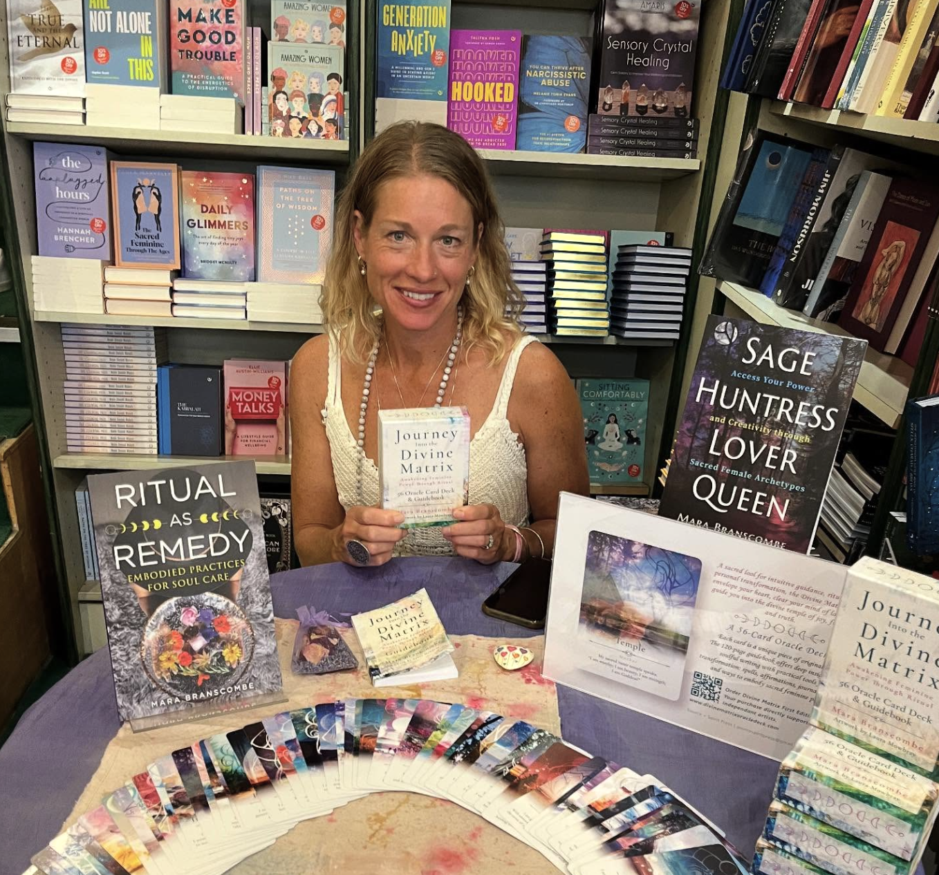 Mara Branscombe with Sage Huntress Lover Queen book, Journey to the Divine Matrix oracle deck, and spiritual wellness books at author event